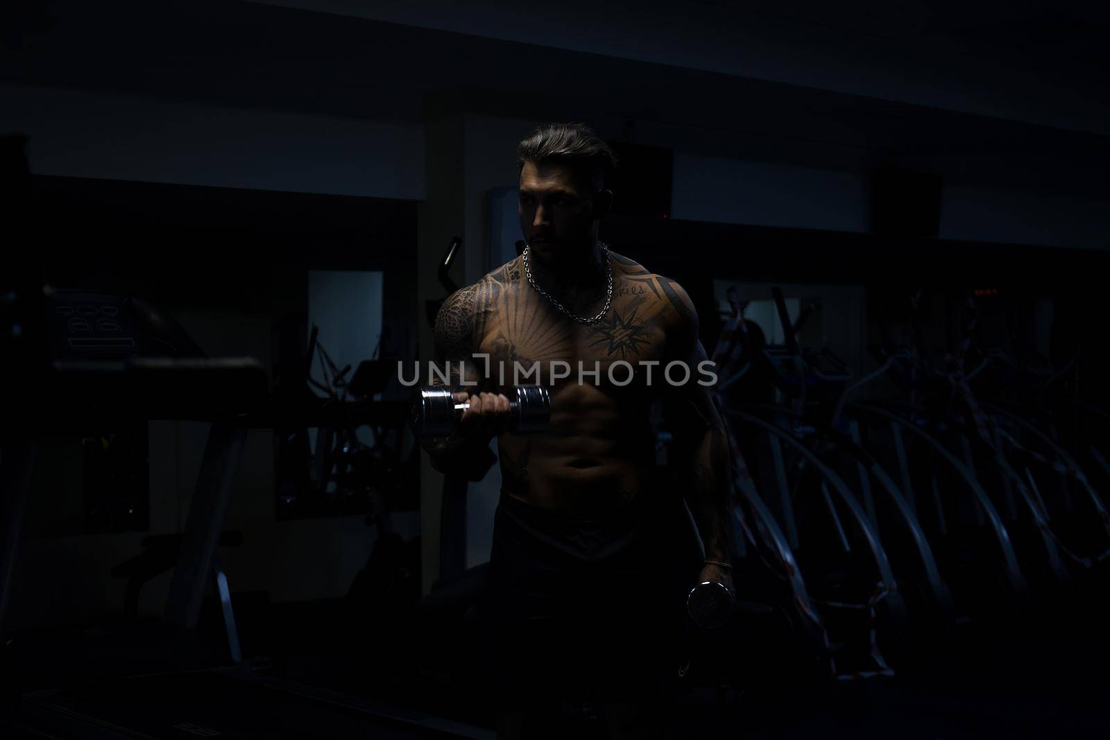 Muscular male athlete with tattooed naked torso standing in dark gym and doing dumbbell curls during workout