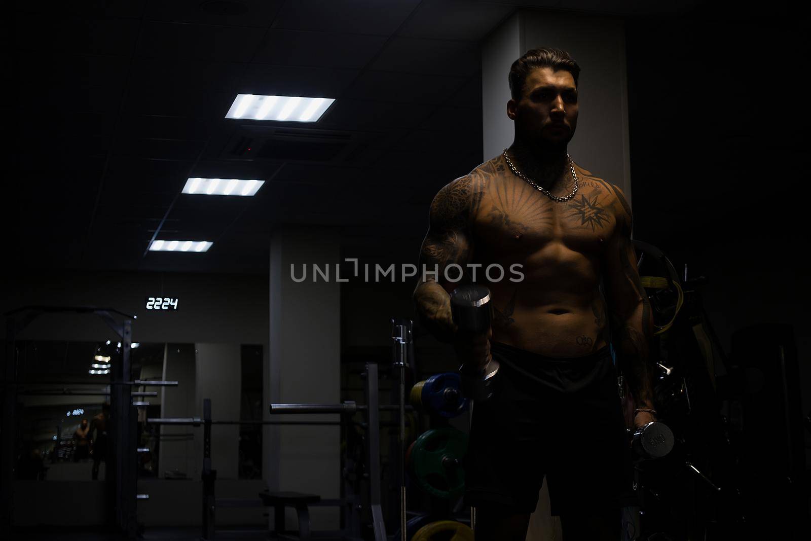 Muscular male athlete with tattooed naked torso standing in dark gym and doing dumbbell curls during workout