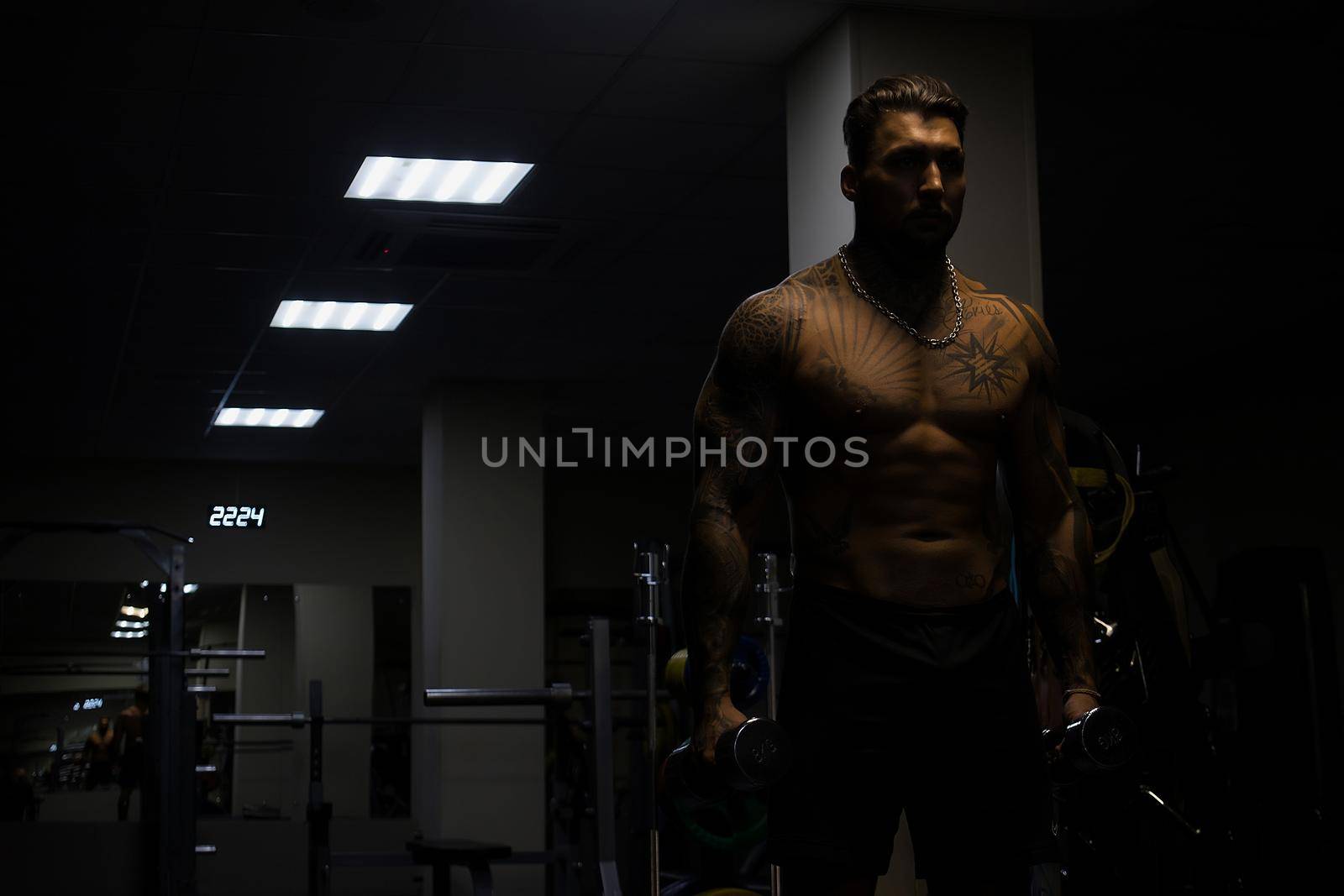 Muscular male athlete with tattooed naked torso standing in dark gym and doing dumbbell curls during workout