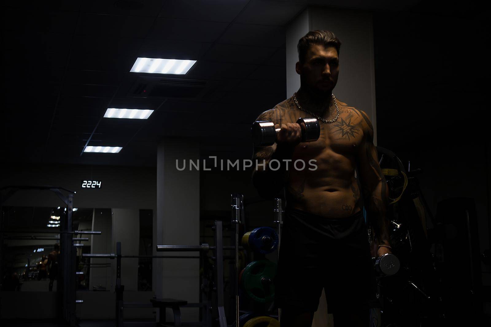 Muscular male athlete with tattooed naked torso standing in dark gym and doing dumbbell curls during workout