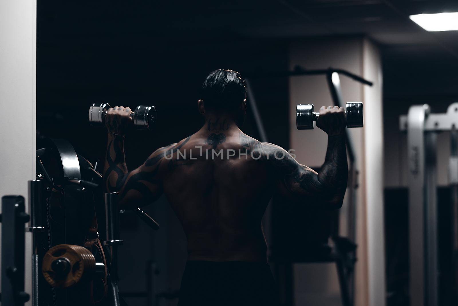 Muscular male athlete with tattooed naked torso standing in dark gym and doing dumbbell curls during workout