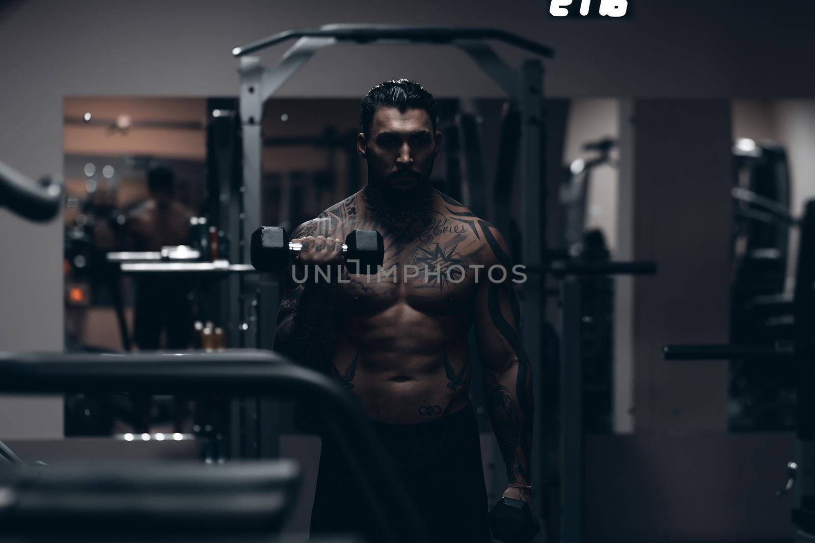 Muscular male athlete with tattooed naked torso standing in dark gym and doing dumbbell curls during workout