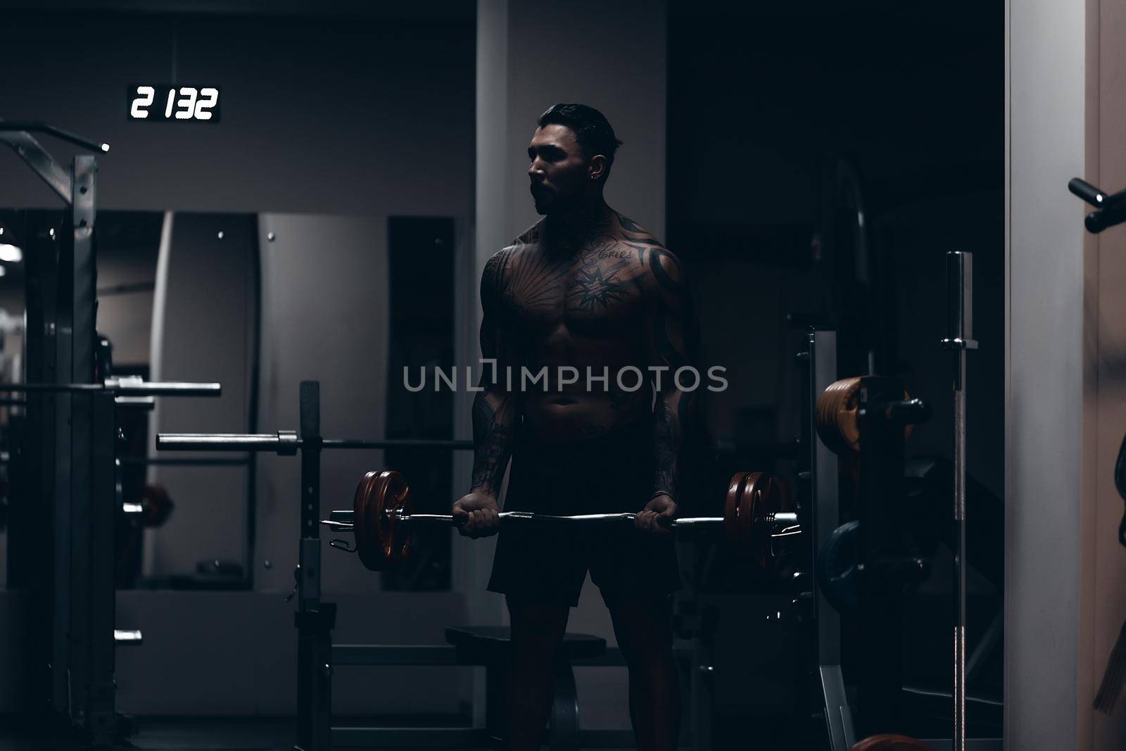 Muscular male athlete with tattooed naked torso standing in dark gym and doing dumbbell curls during workout