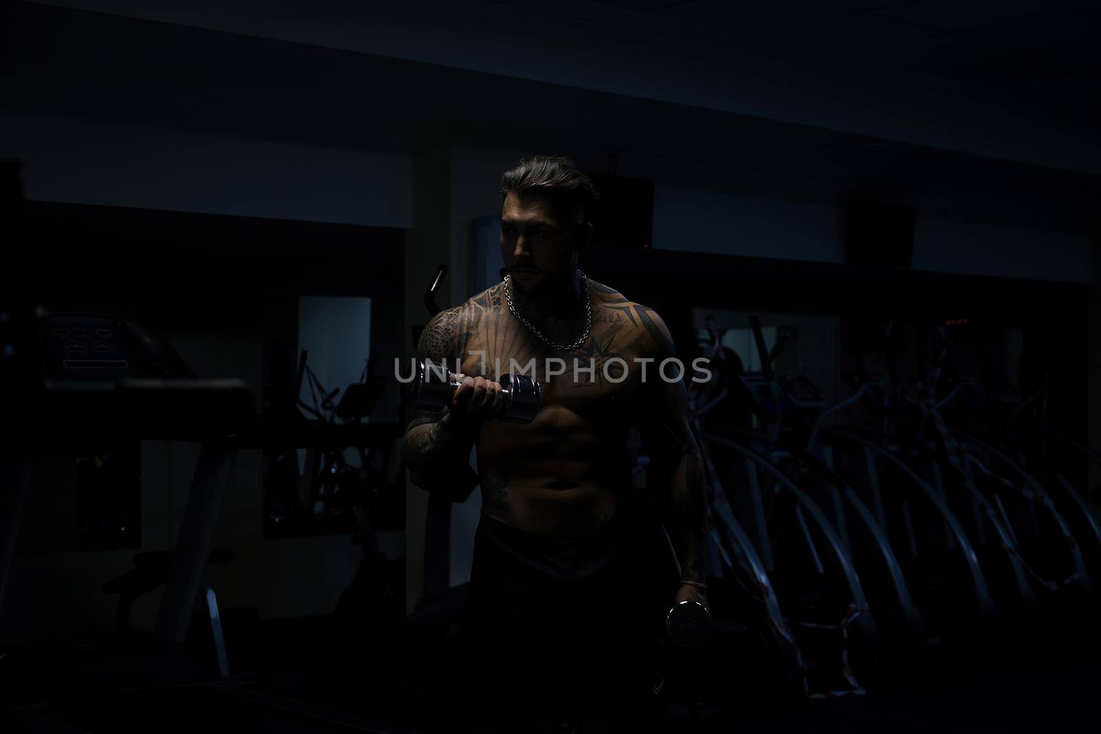 Muscular male athlete with tattooed naked torso standing in dark gym and doing dumbbell curls during workout