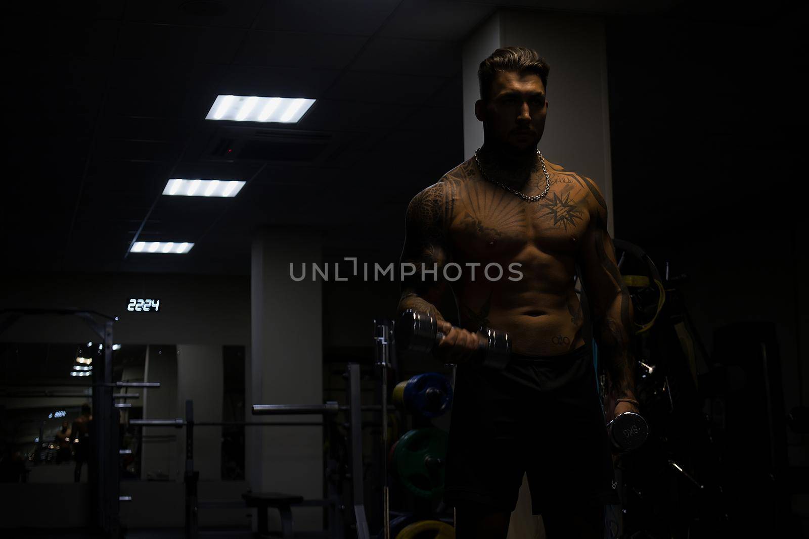 Muscular male athlete with tattooed naked torso standing in dark gym and doing dumbbell curls during workout