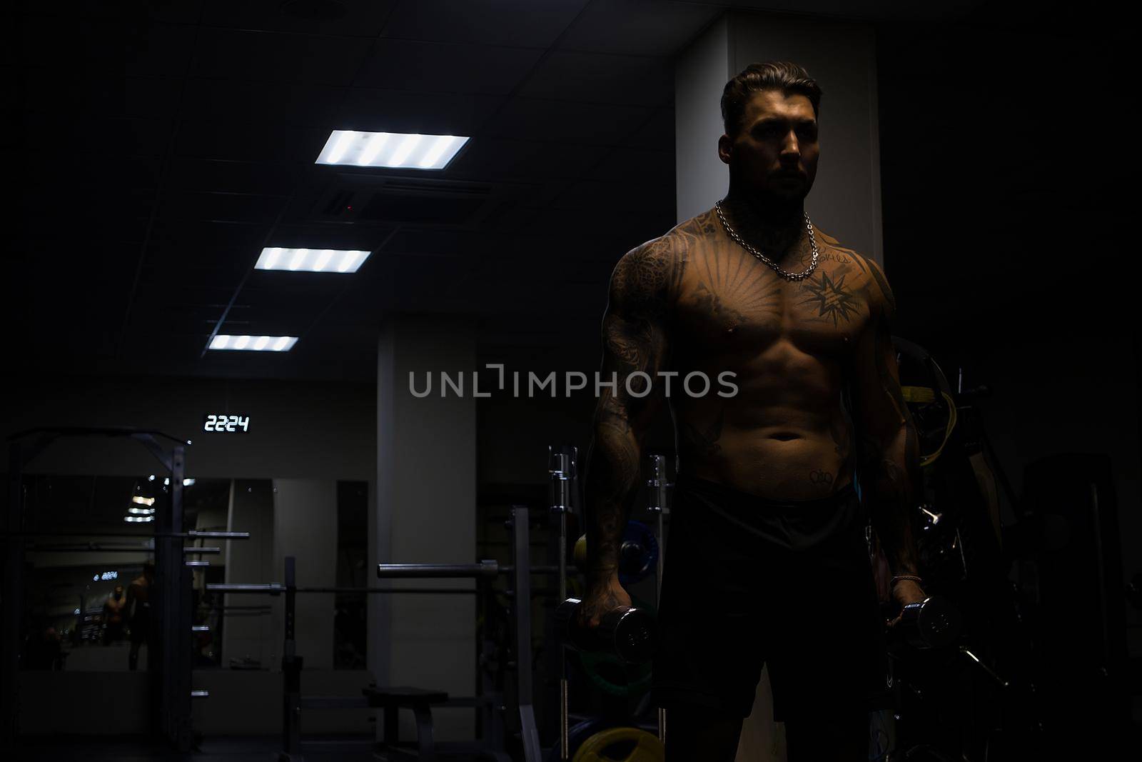 Muscular male athlete with tattooed naked torso standing in dark gym and doing dumbbell curls during workout