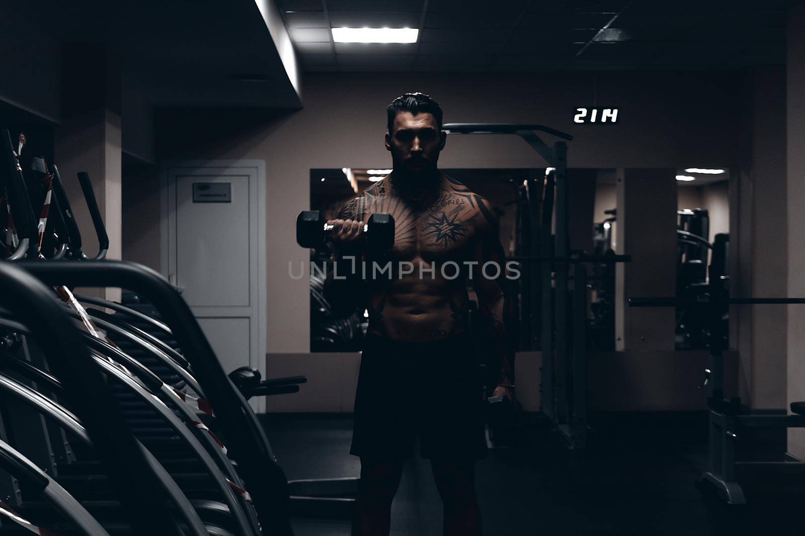 Muscular male athlete with tattooed naked torso standing in dark gym and doing dumbbell curls during workout