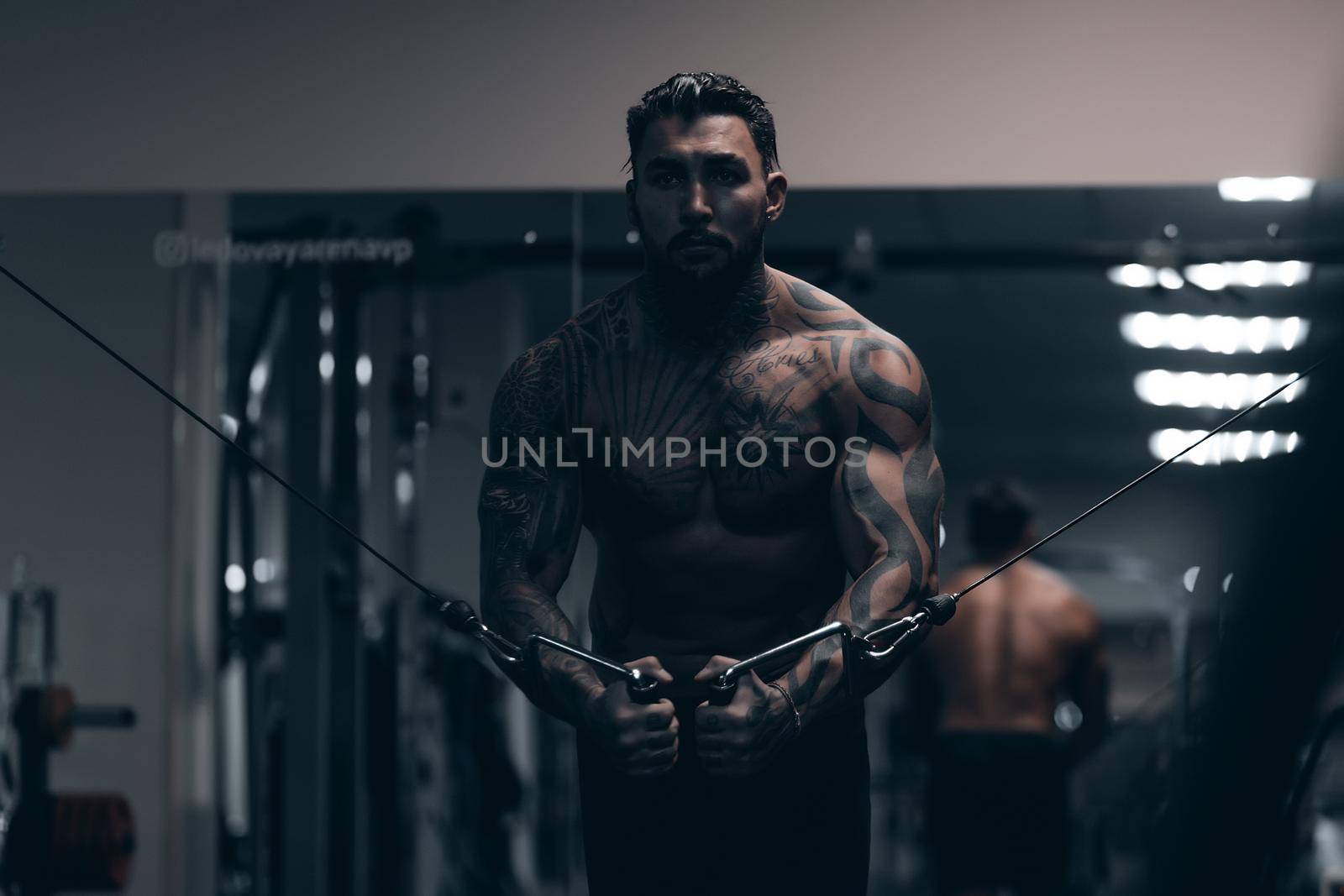 Muscular male athlete with tattooed naked torso standing in dark gym and doing dumbbell curls during workout