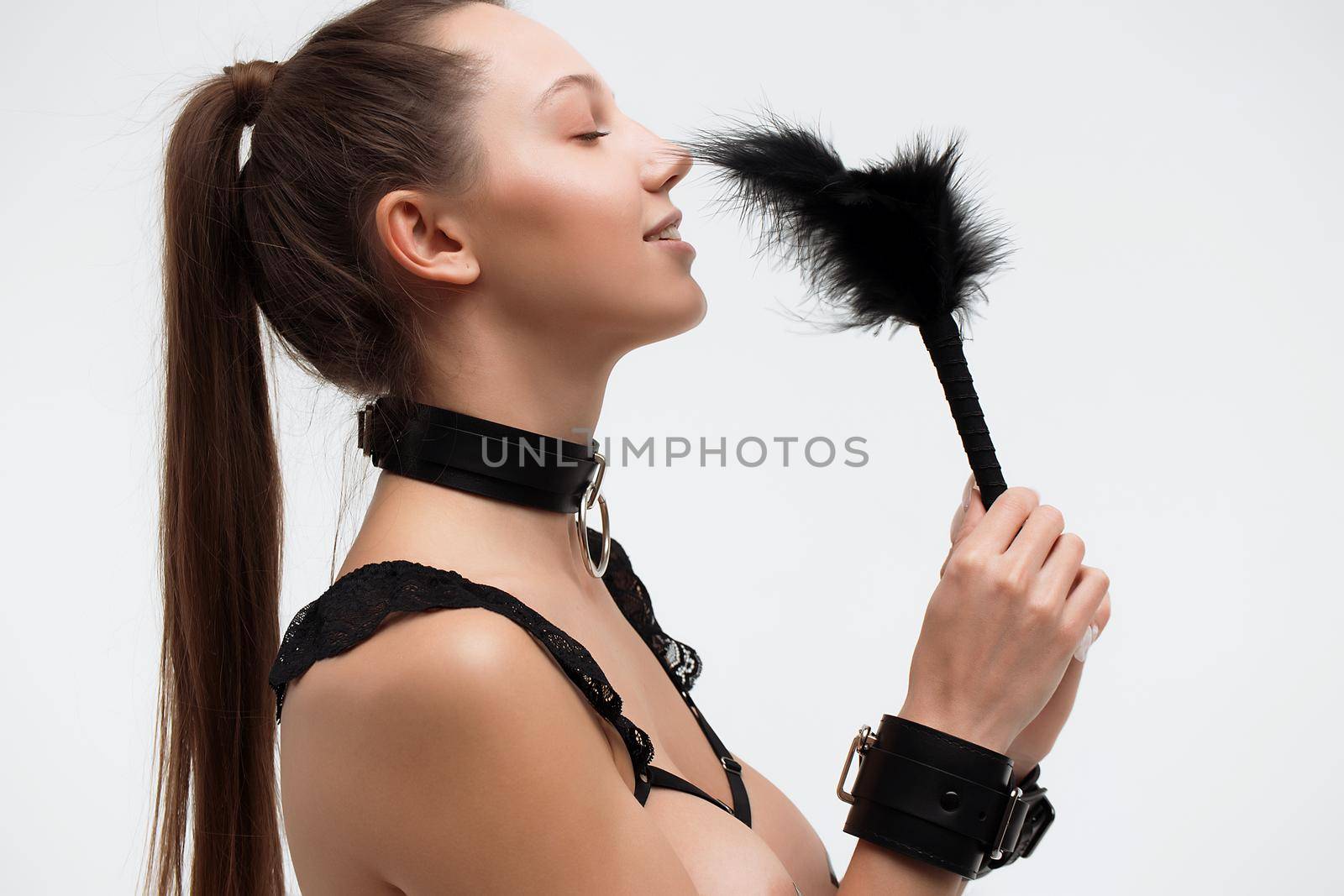 Side view of seductive young woman in underwear and collar smiling and tickling nose with feather against gray background