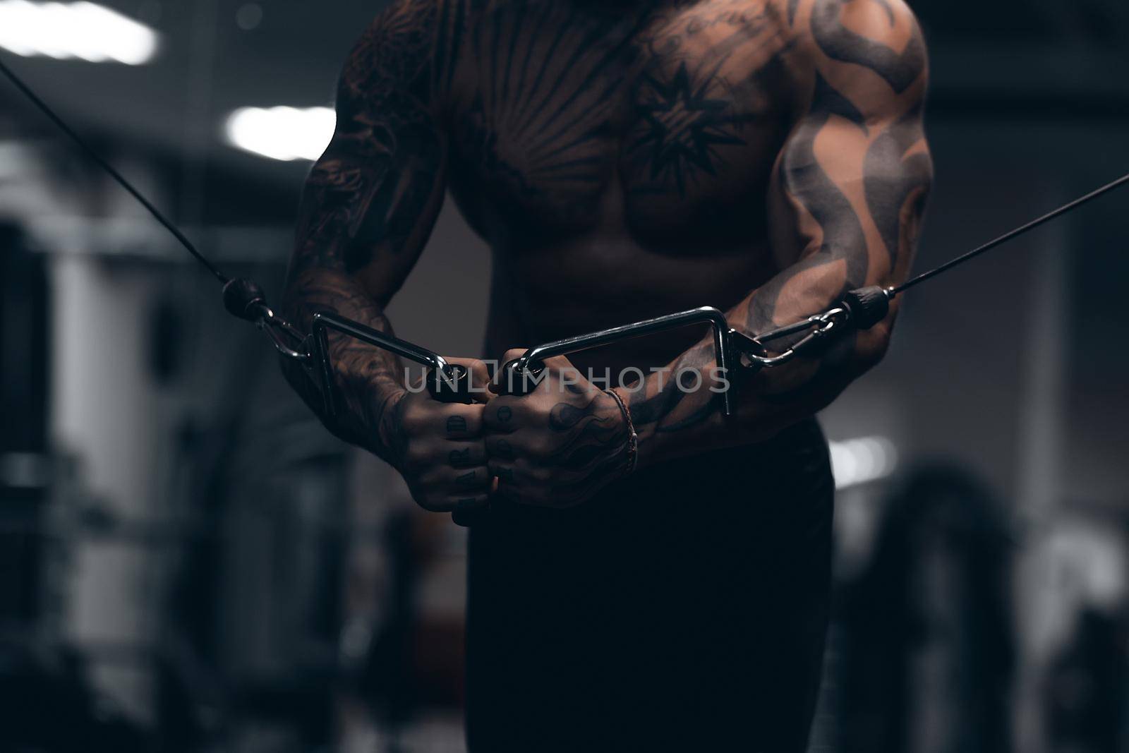 Muscular male athlete with tattooed naked torso standing in dark gym and doing dumbbell curls during workout