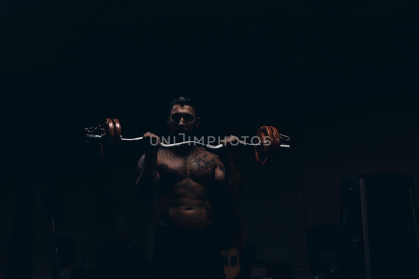 Muscular male athlete with tattooed naked torso standing in dark gym and doing dumbbell curls during workout