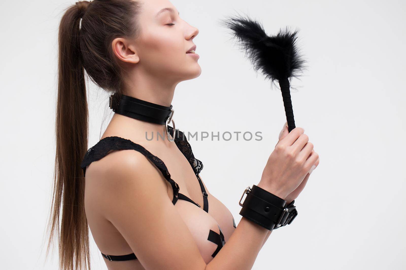 Side view of seductive young woman in underwear and collar smiling and tickling nose with feather against gray background