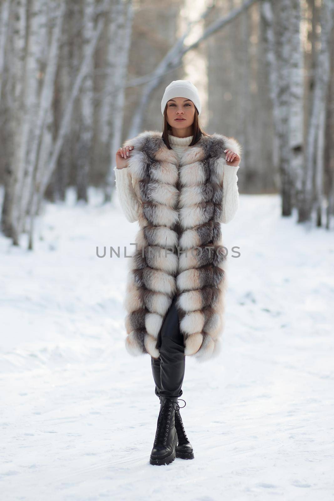 Gorgeous young brunette in fur vest and white knitwear looking confidently away against trees in winter woods