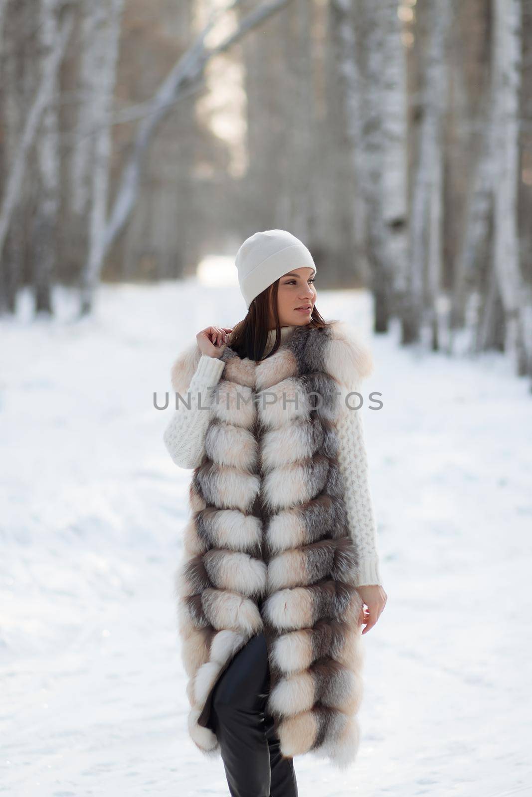 Gorgeous young brunette in fur vest and white knitwear looking confidently away against trees in winter woods