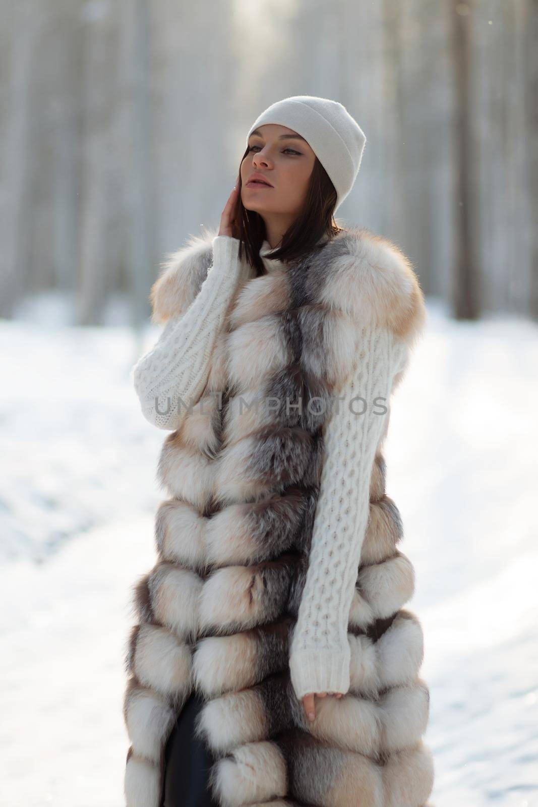 Gorgeous young brunette in fur vest and white knitwear looking confidently away against trees in winter woods