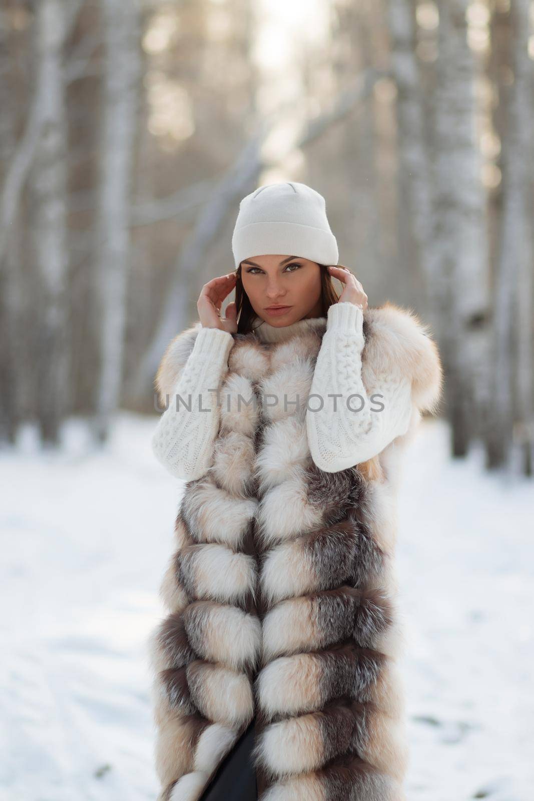 Gorgeous young brunette in fur vest and white knitwear looking confidently away against trees in winter woods