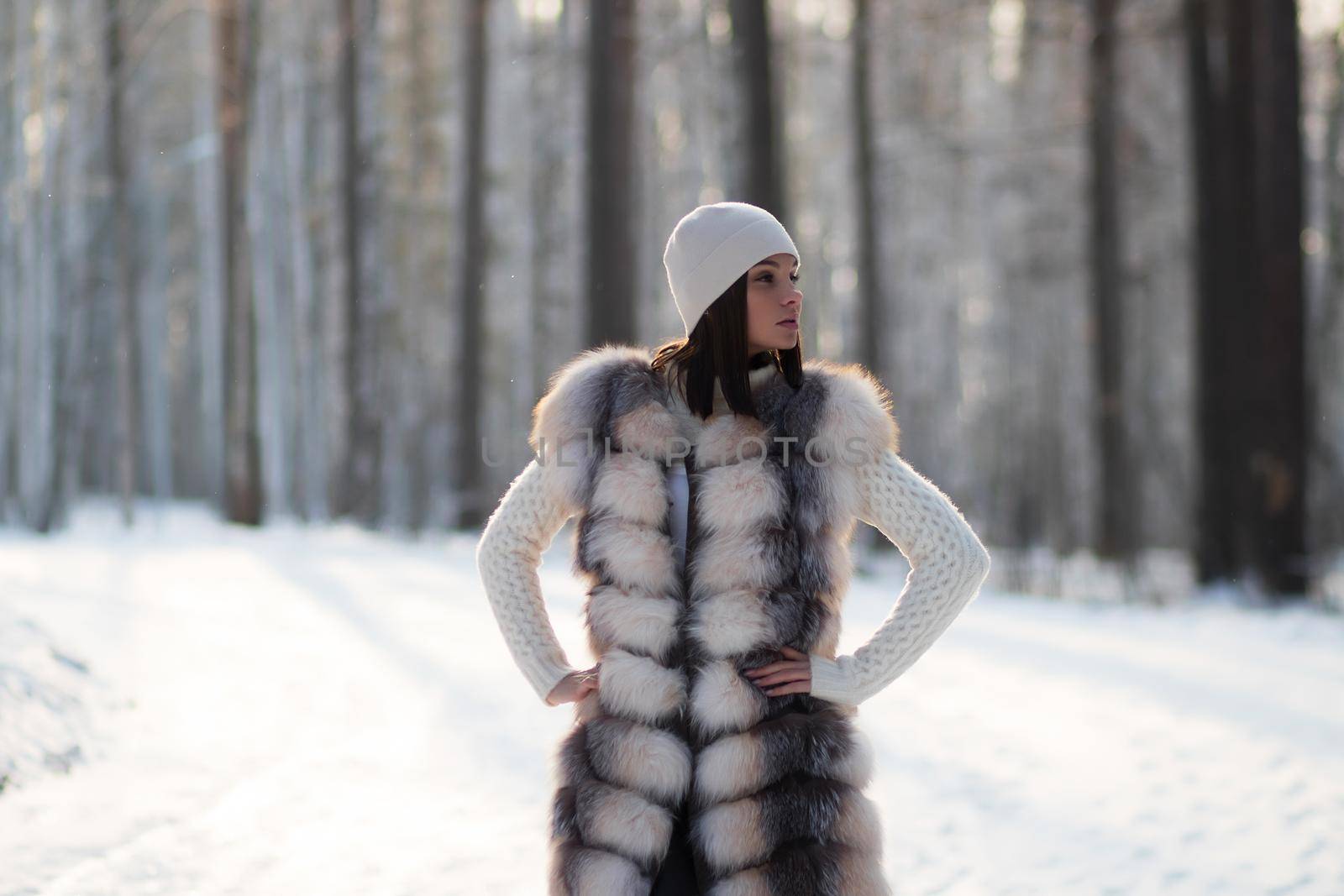 Gorgeous young brunette in fur vest and white knitwear looking confidently away against trees in winter woods