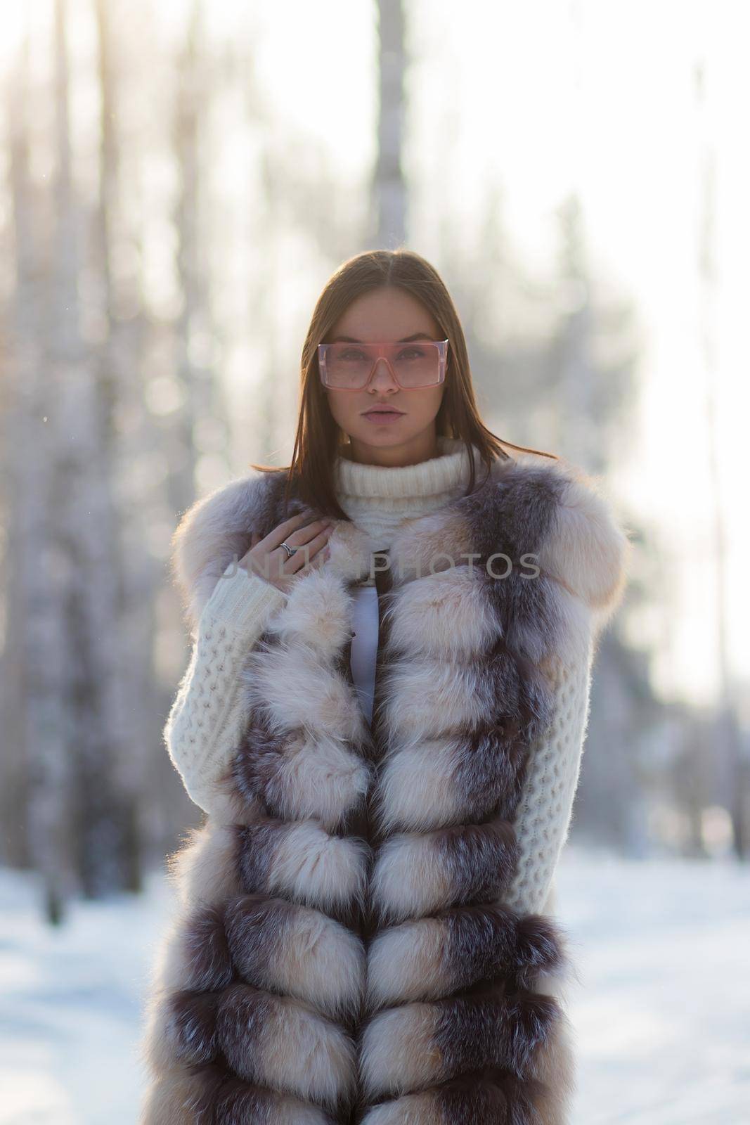 Gorgeous young brunette in fur vest and white knitwear looking confidently away against trees in winter woods