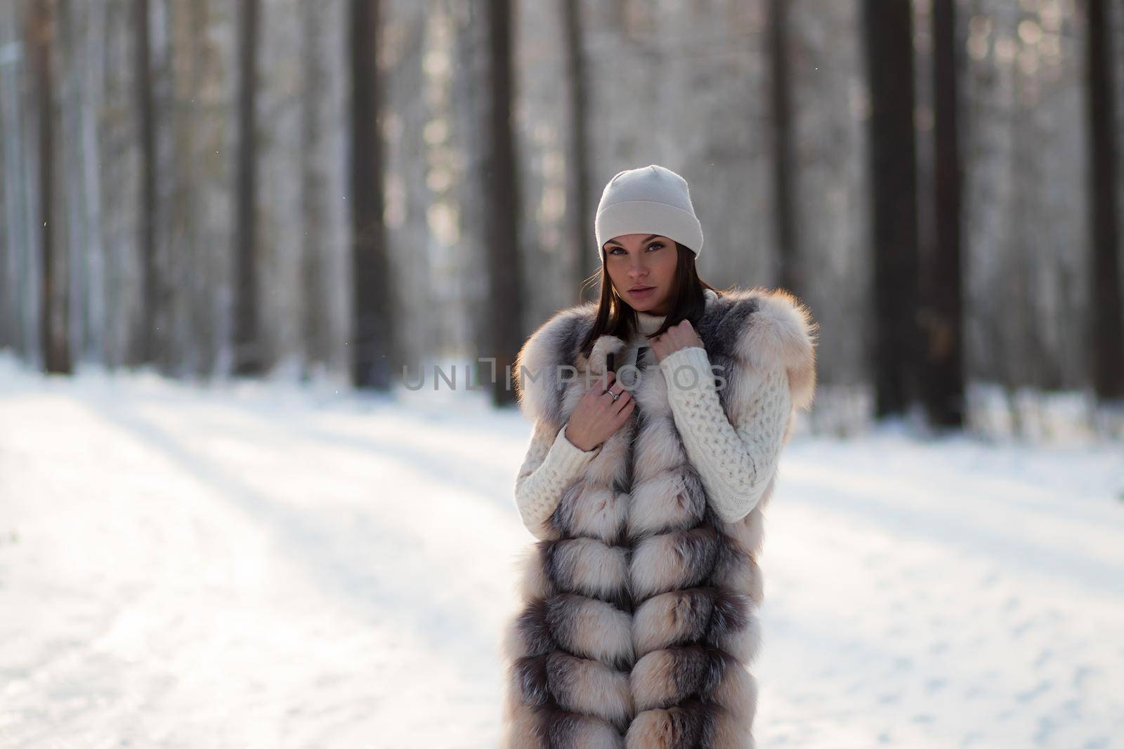 Gorgeous young brunette in fur vest and white knitwear looking confidently away against trees in winter woods