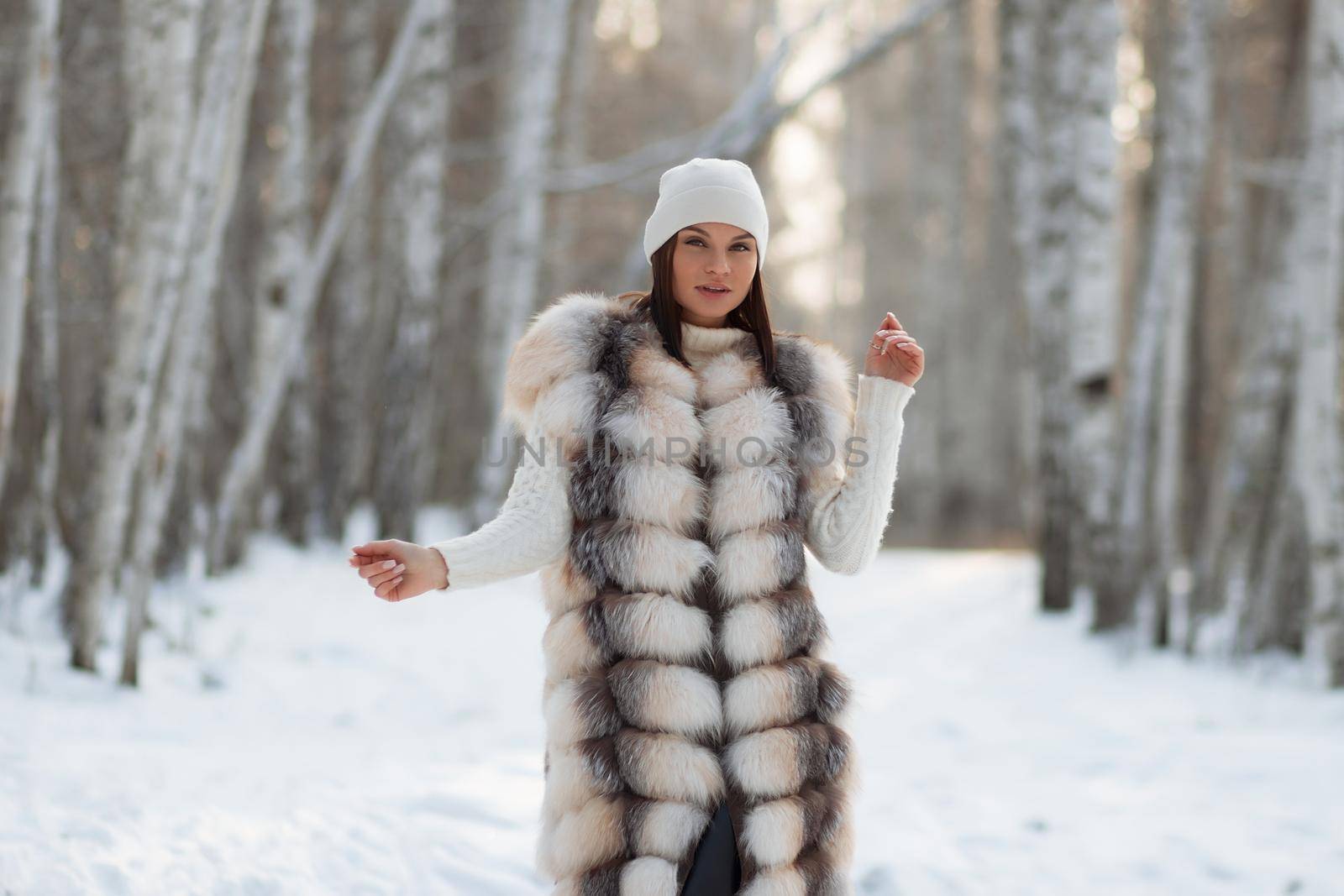 Gorgeous young brunette in fur vest and white knitwear looking confidently away against trees in winter woods