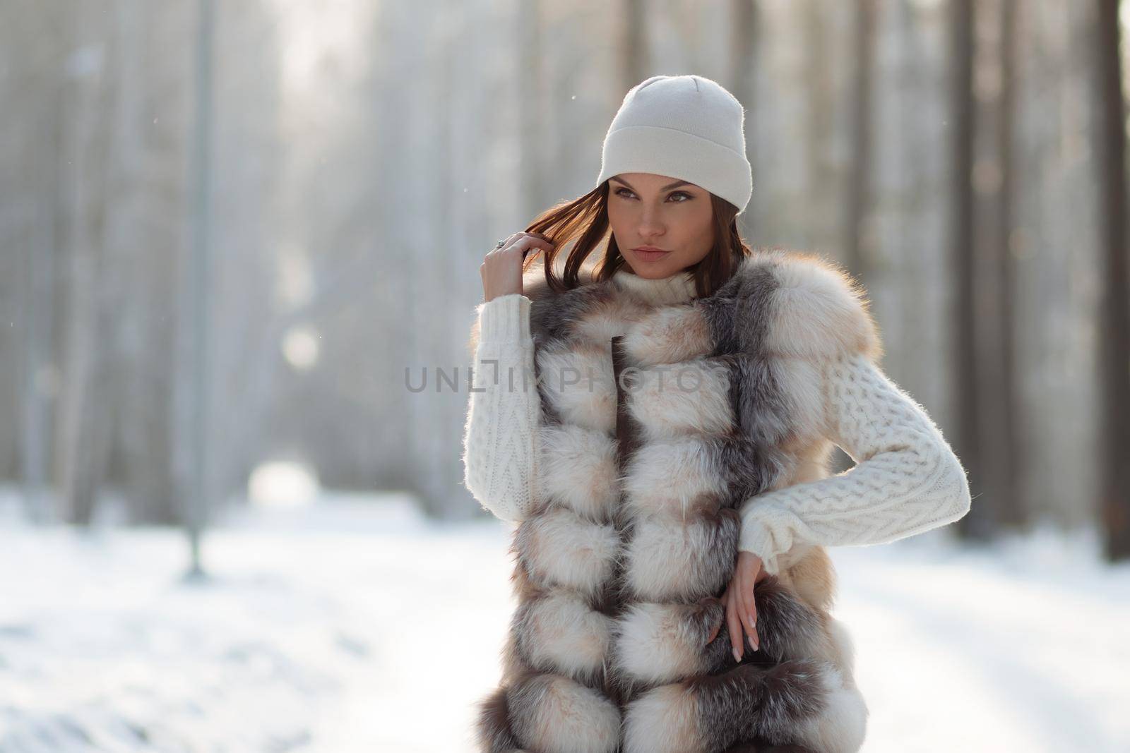 Gorgeous young brunette in fur vest and white knitwear looking confidently away against trees in winter woods