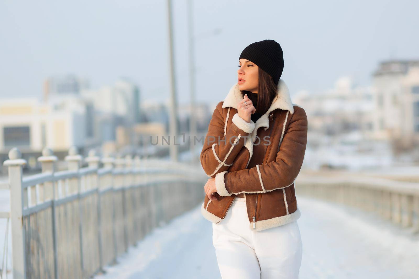 Alluring woman wearing brown sheepskin jacket with hat and looking at camera standing on snowy city bridge