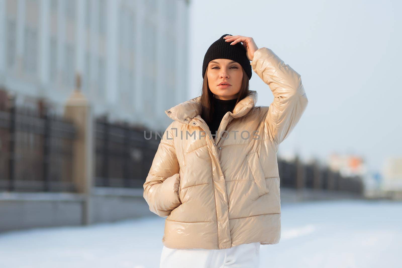 Alluring woman wearing brown sheepskin jacket with hat and looking at camera standing on snowy city bridge