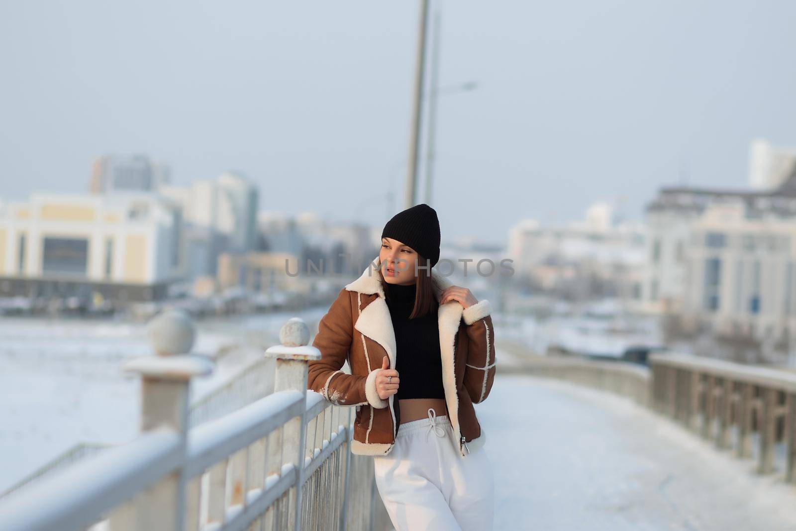 Alluring woman wearing brown sheepskin jacket with hat and looking at camera standing on snowy city bridge