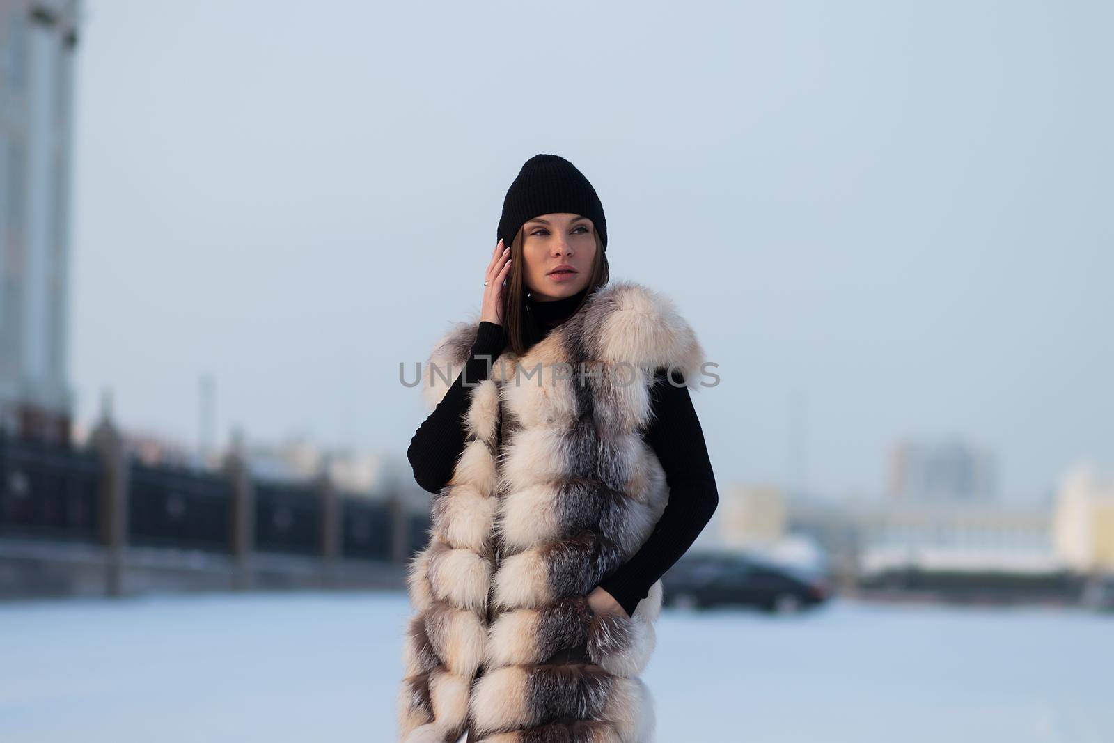 Alluring woman wearing brown sheepskin jacket with hat and looking at camera standing on snowy city bridge