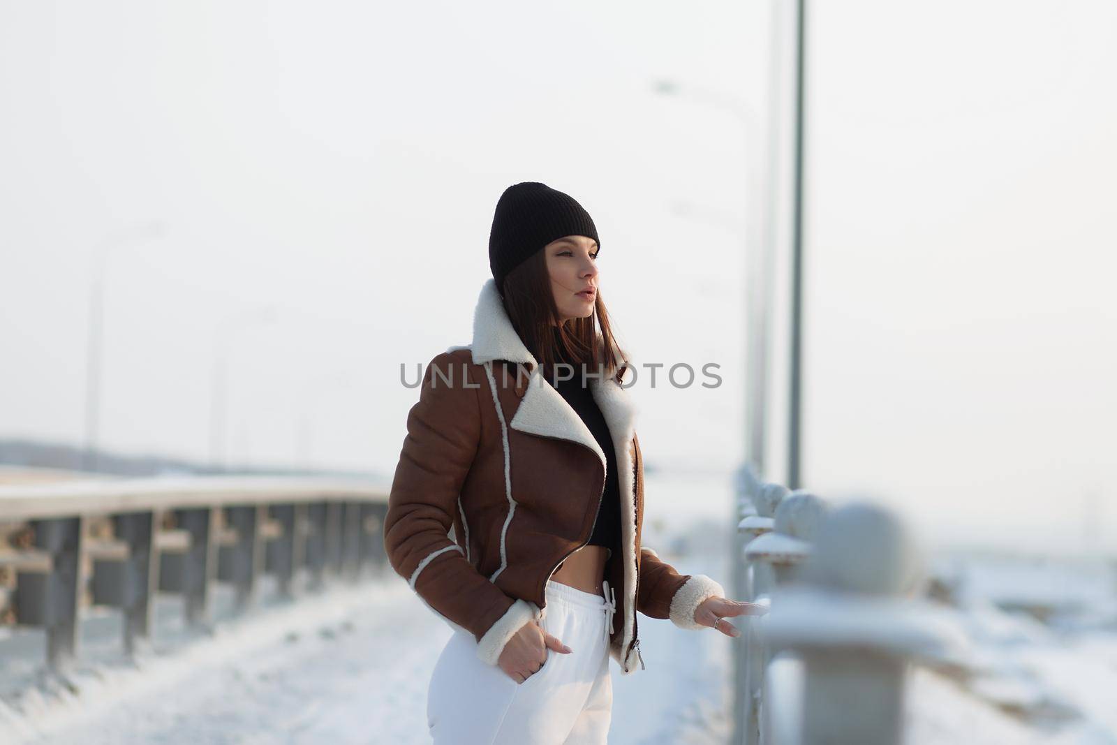 Alluring woman wearing brown sheepskin jacket with hat and looking at camera standing on snowy city bridge