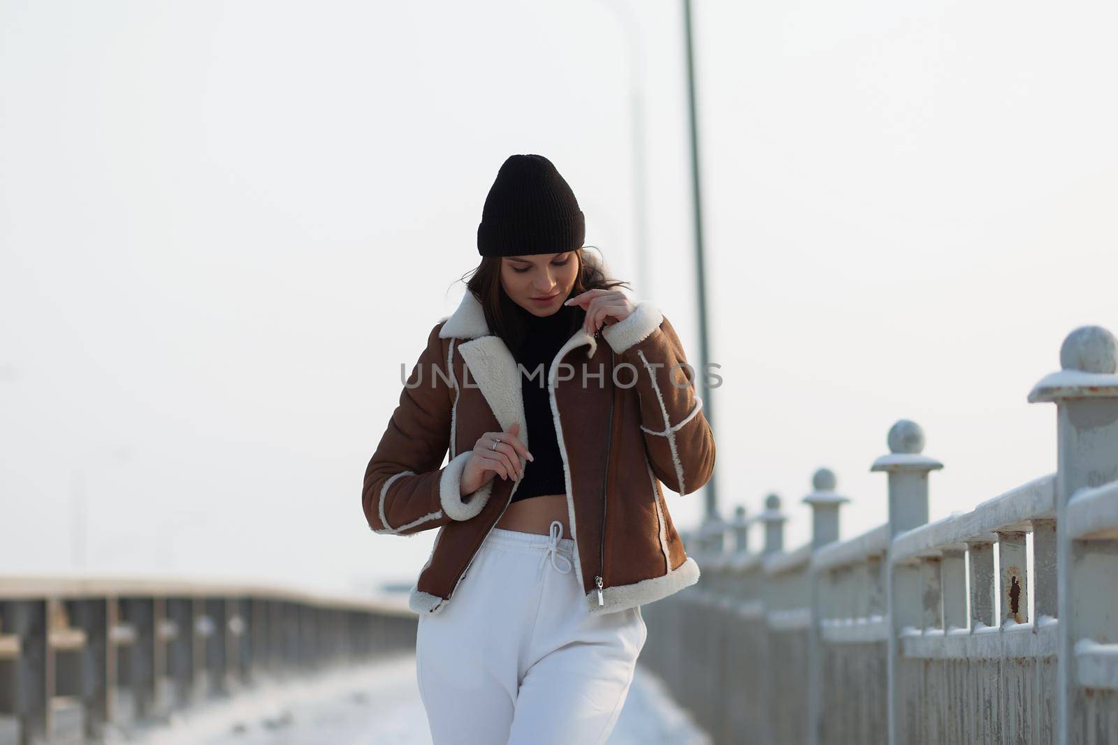 Alluring woman wearing brown sheepskin jacket with hat and looking at camera standing on snowy city bridge