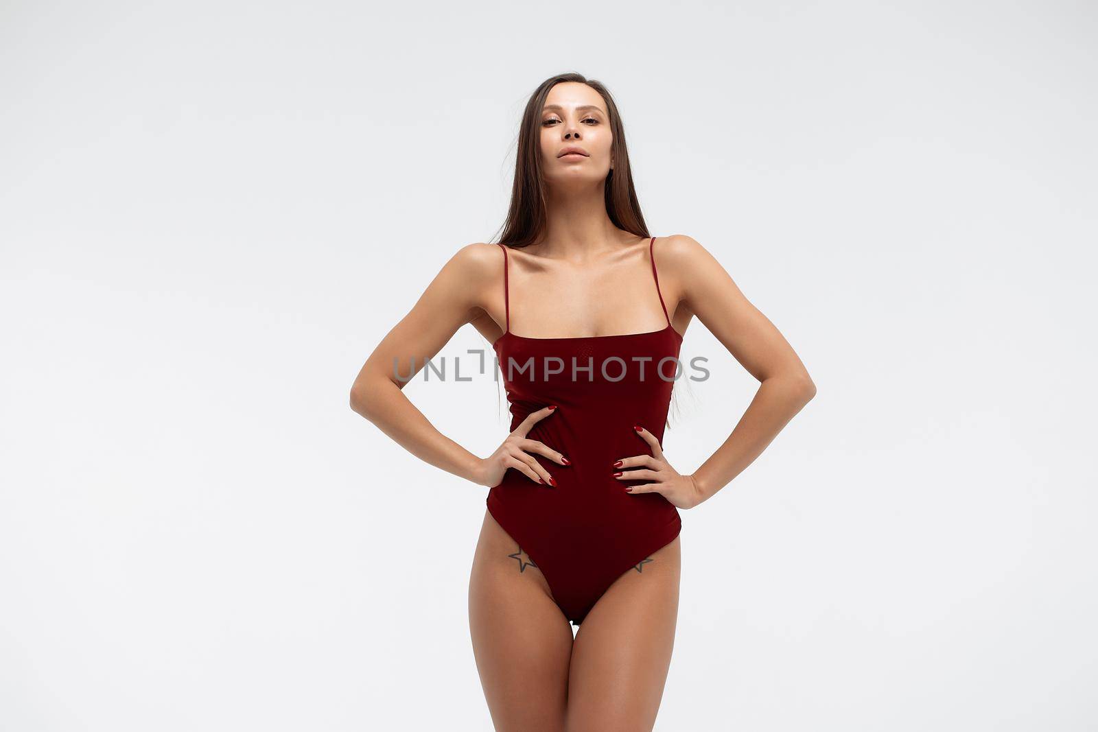 Sexy young female wearing red bodysuit and high heeled shoes standing on white background in studio and looking at camera