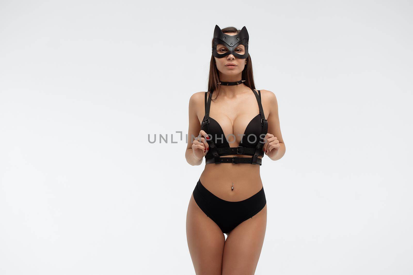 Slim seductive female wearing black mask and sexy underwear standing with hands on waist on white background and looking at camera