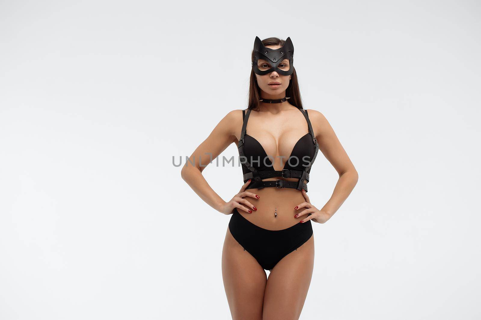 Slim seductive female wearing black mask and sexy underwear standing with hands on waist on white background and looking at camera