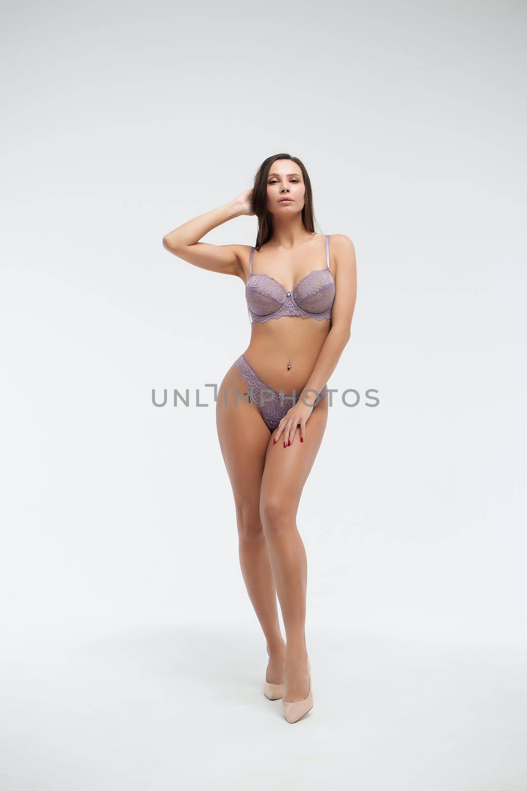 Coquette female model in lace purple bra and panties standing on white background in studio and touching hair while looking at camera