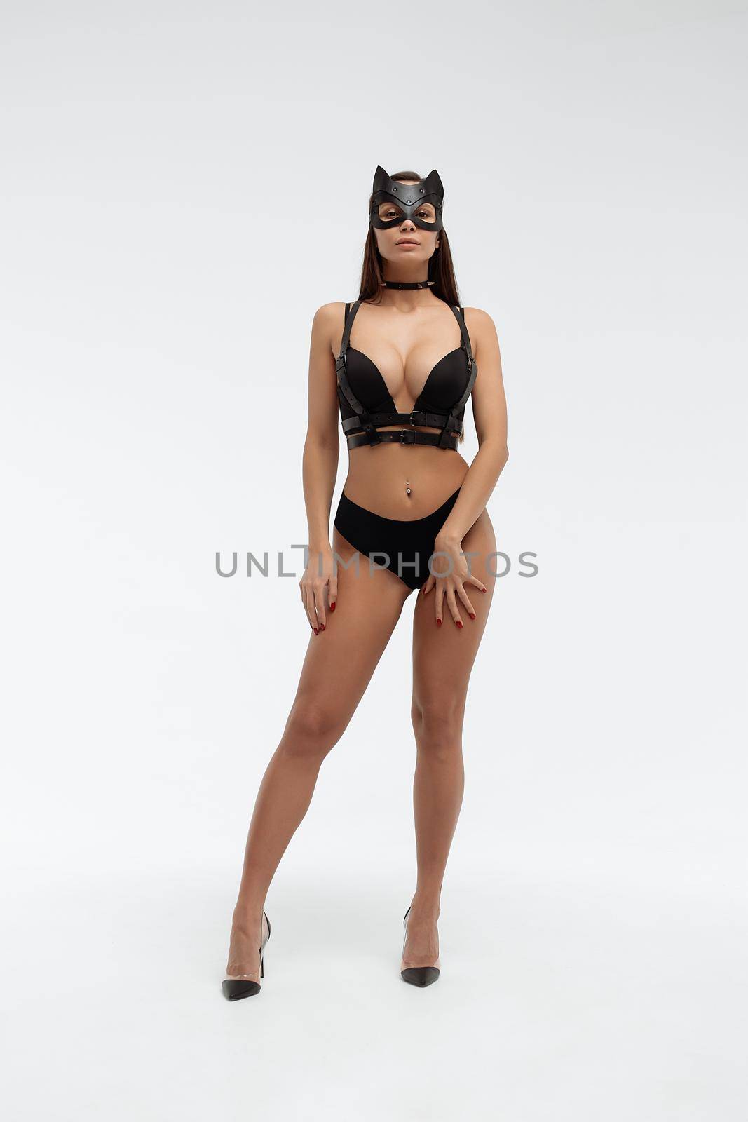 Slim seductive female wearing black mask and sexy underwear standing with hands on waist on white background and looking at camera