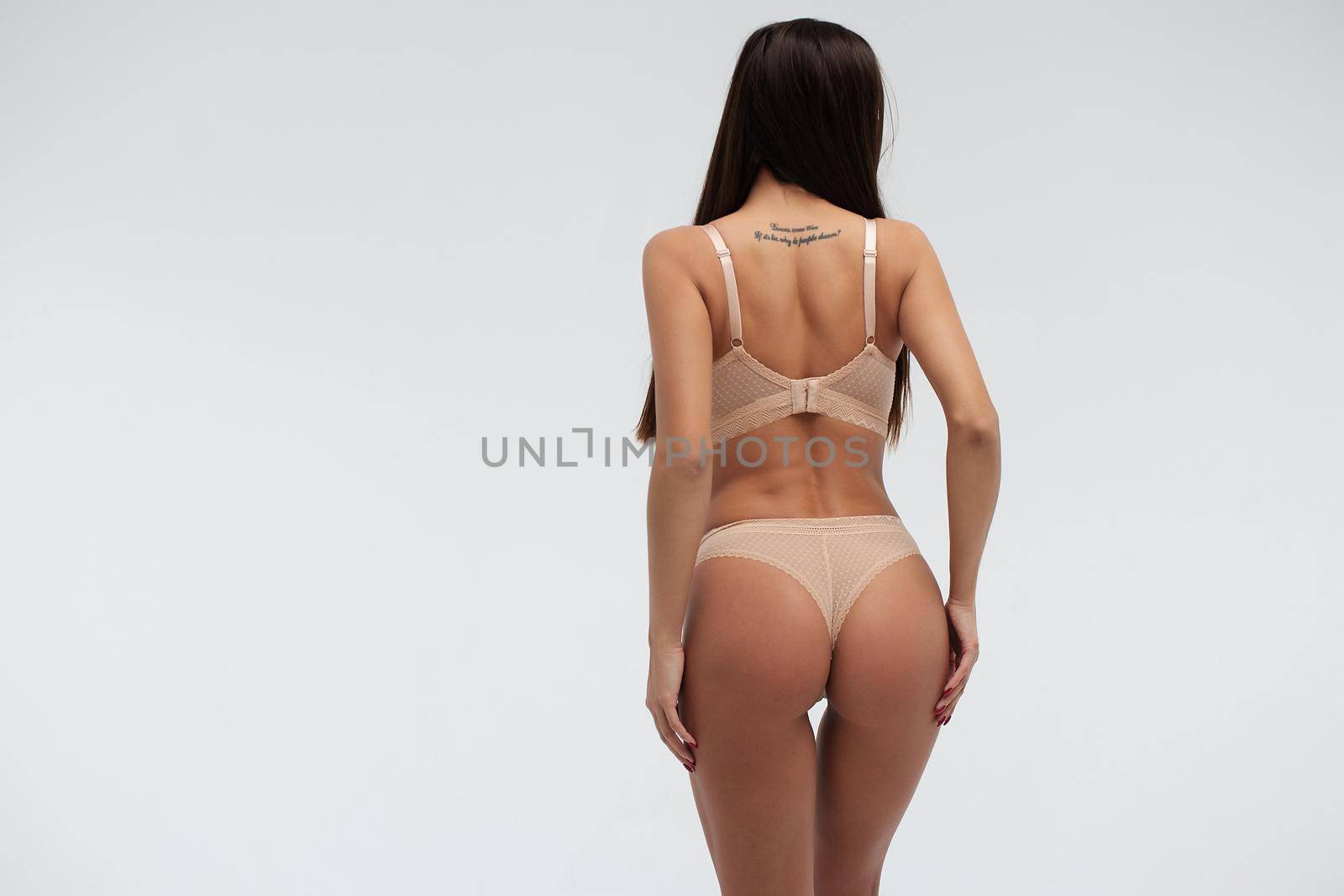 Back view of unrecognizable slim female wearing beige lingerie standing on white background in studio