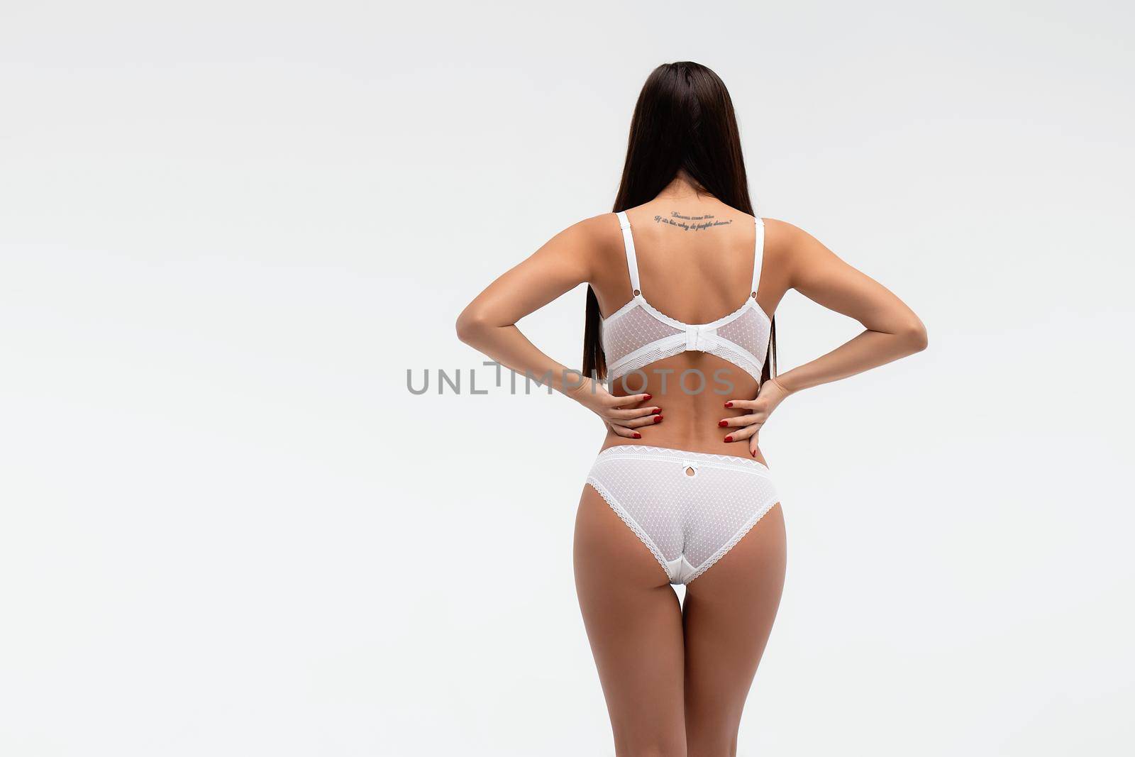 Back view of unrecognizable slim female wearing white lingerie standing on white background in studio