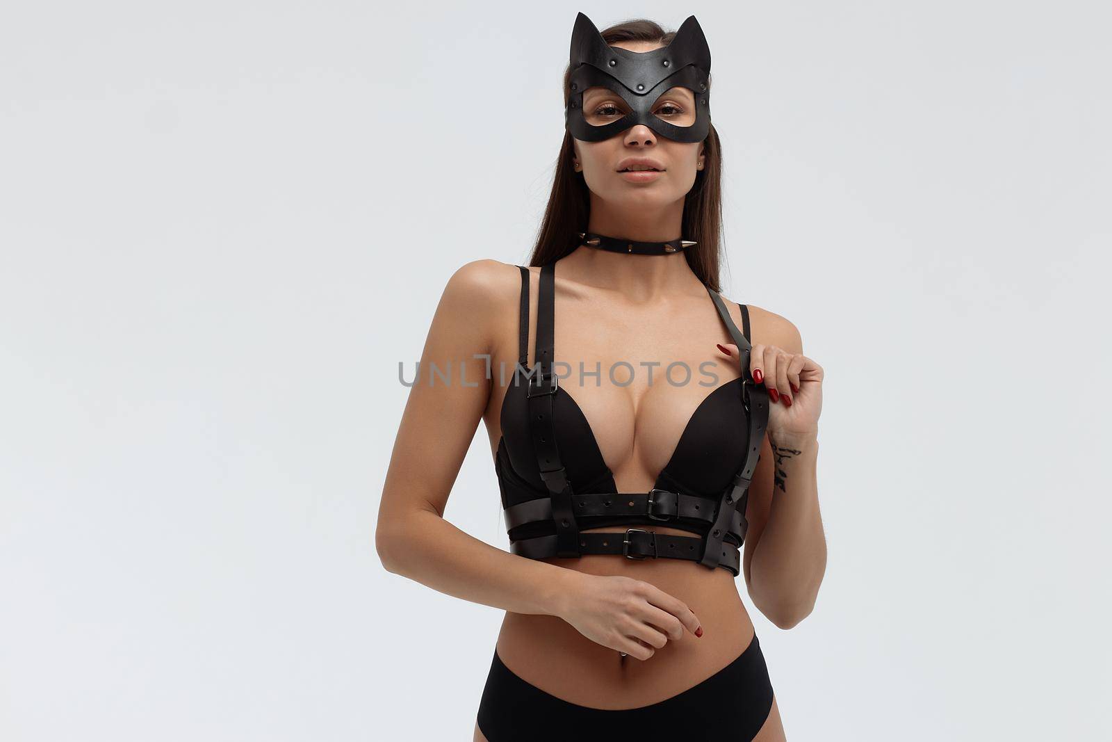 Slim seductive female wearing black mask and sexy underwear standing with hands on waist on white background and looking at camera