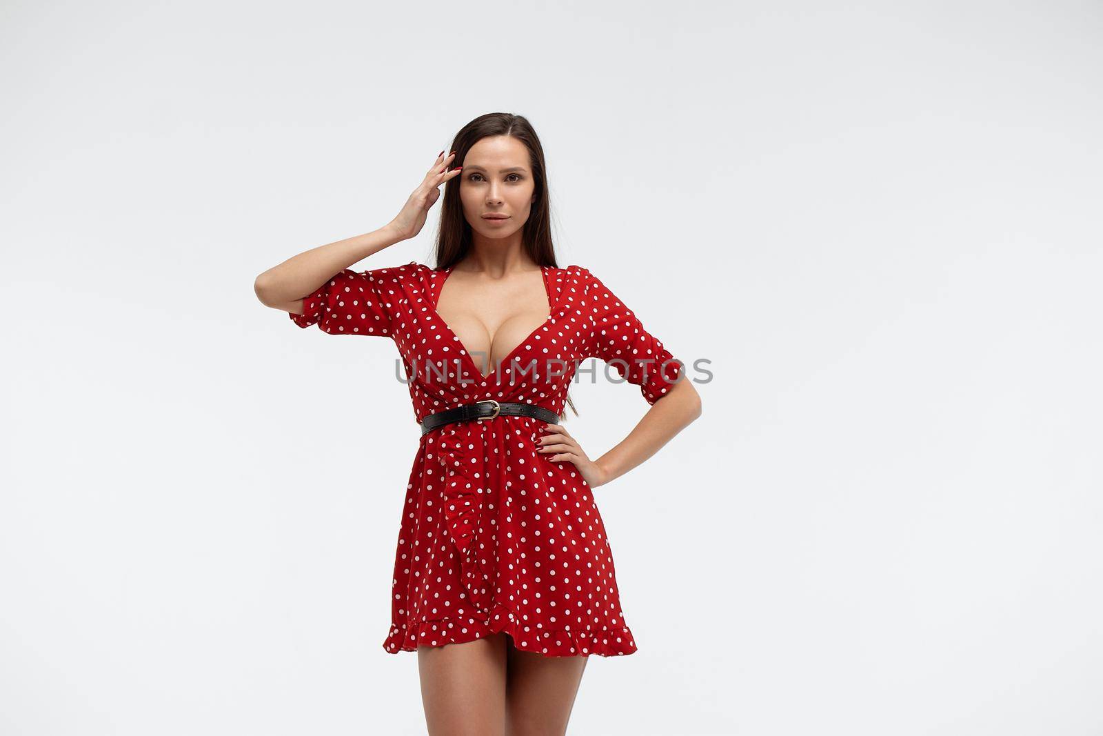 Sexy female wearing red short polka dot dress standing on white background in studio and touching hair while looking at camera