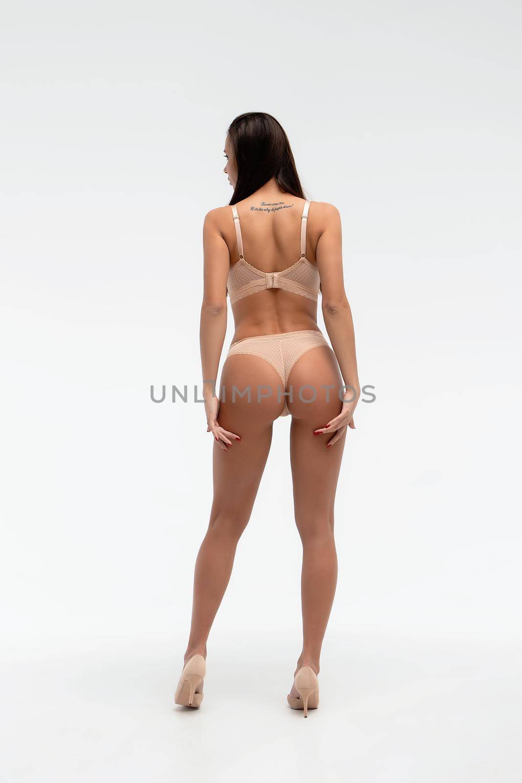 Back view of unrecognizable slim female wearing beige lingerie standing on white background in studio