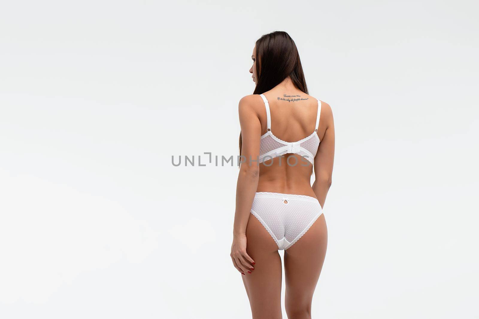 Back view of unrecognizable slim female wearing white lingerie standing on white background in studio