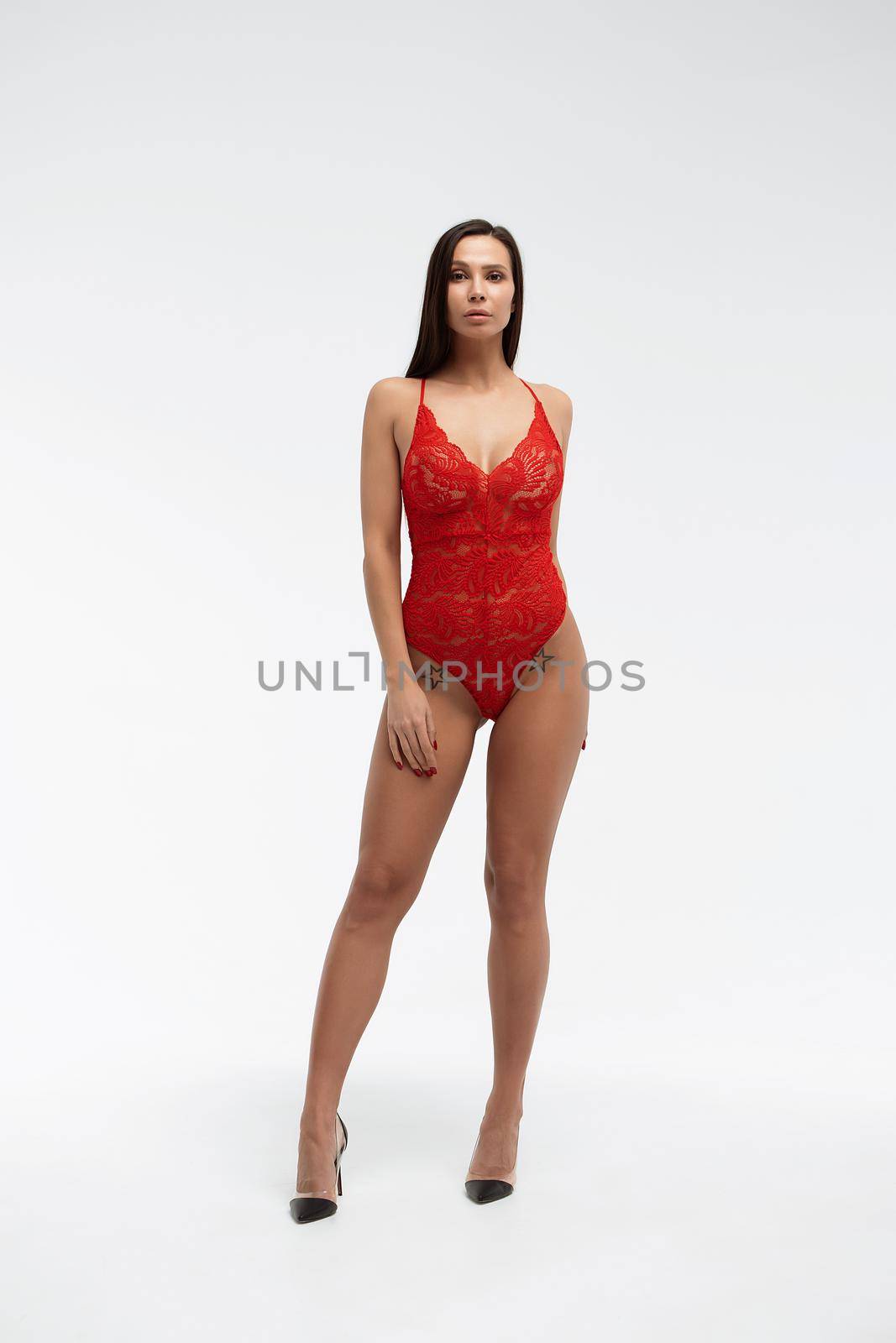 Sexual female with perfect body in red lace bodysuit standing with folded arms on white background and looking at camera