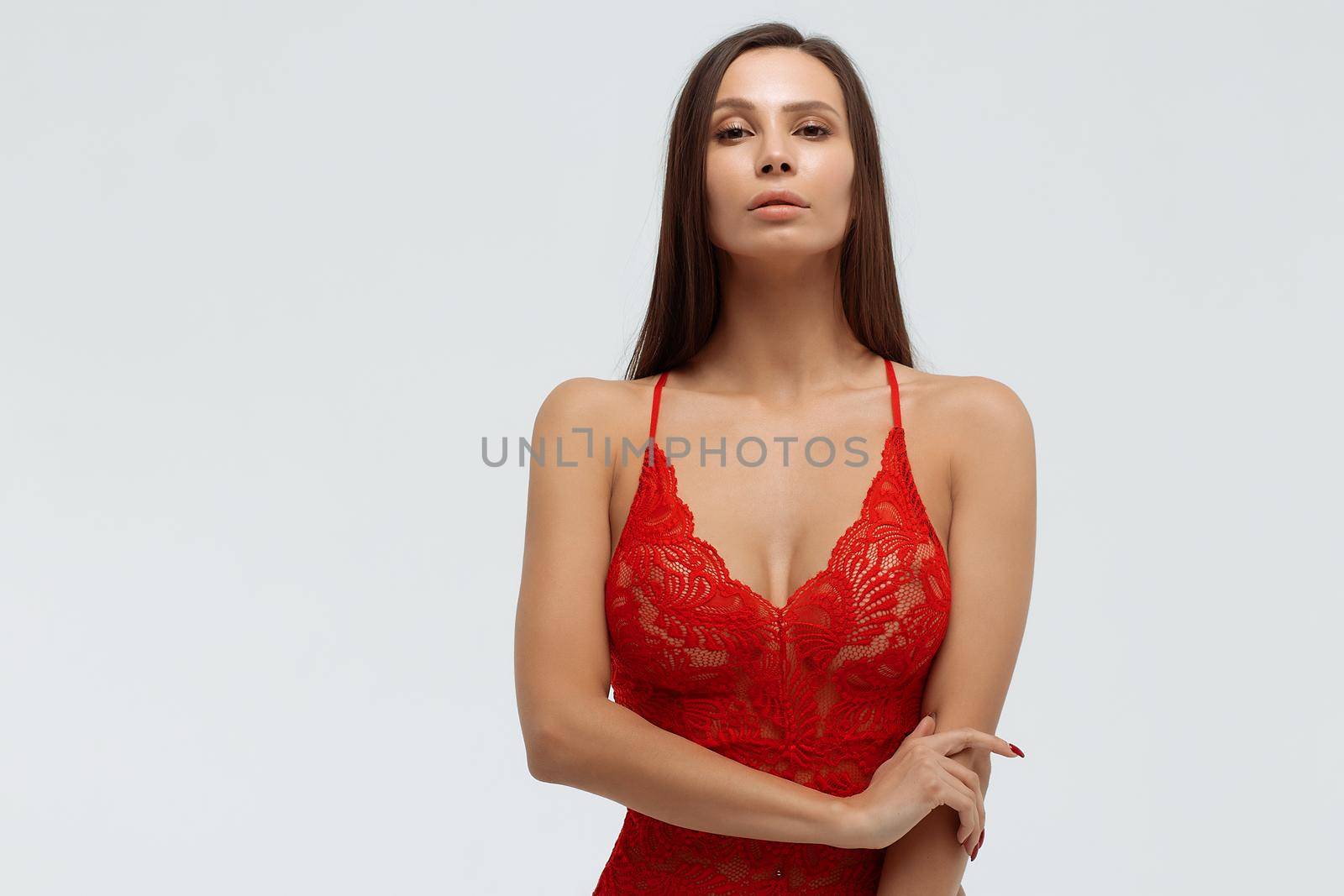 Sexual female with perfect body in red lace bodysuit standing with folded arms on white background and looking at camera