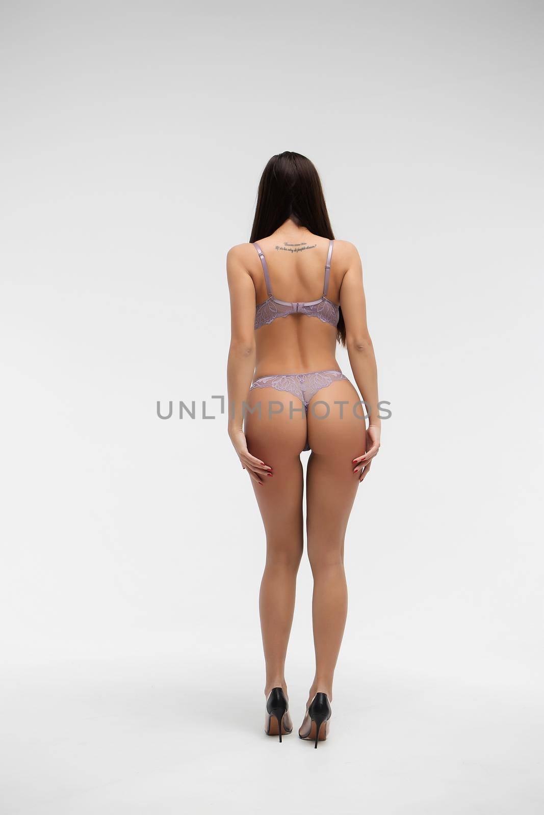 Back view of unrecognizable slim female wearing purple lingerie standing on white background in studio