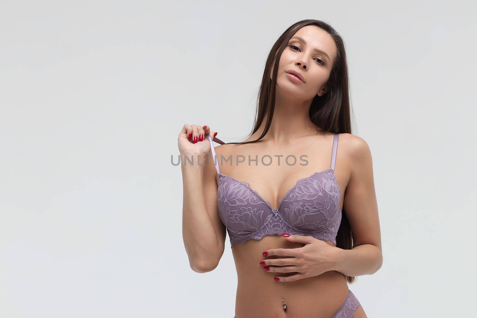 Coquette female model in lace purple bra and panties standing on white background in studio and touching hair while looking at camera