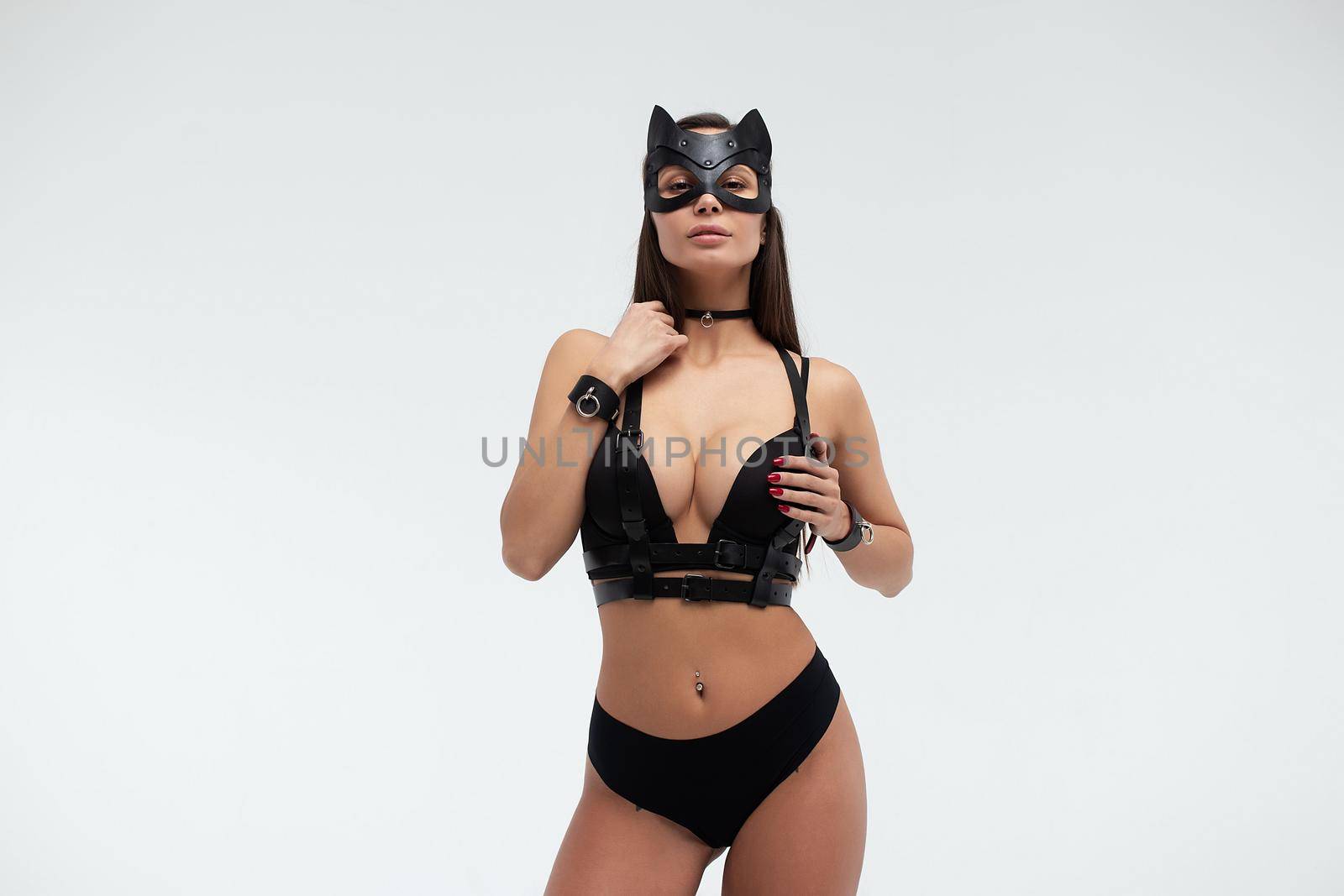 Slim seductive female wearing black mask and sexy underwear standing with hands on waist on white background and looking at camera