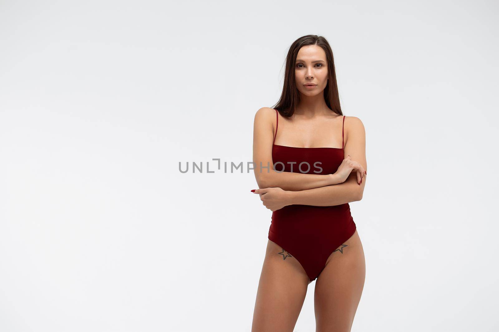 Sexy young female wearing red bodysuit and high heeled shoes standing on white background in studio and looking at camera