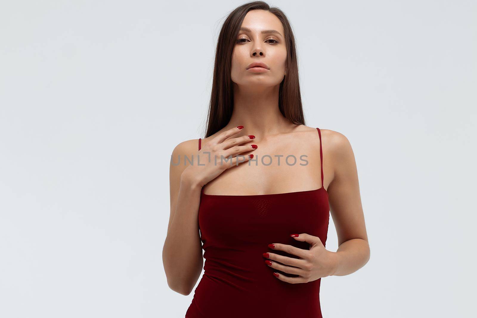 Sexy young female wearing red bodysuit and high heeled shoes standing on white background in studio and looking at camera