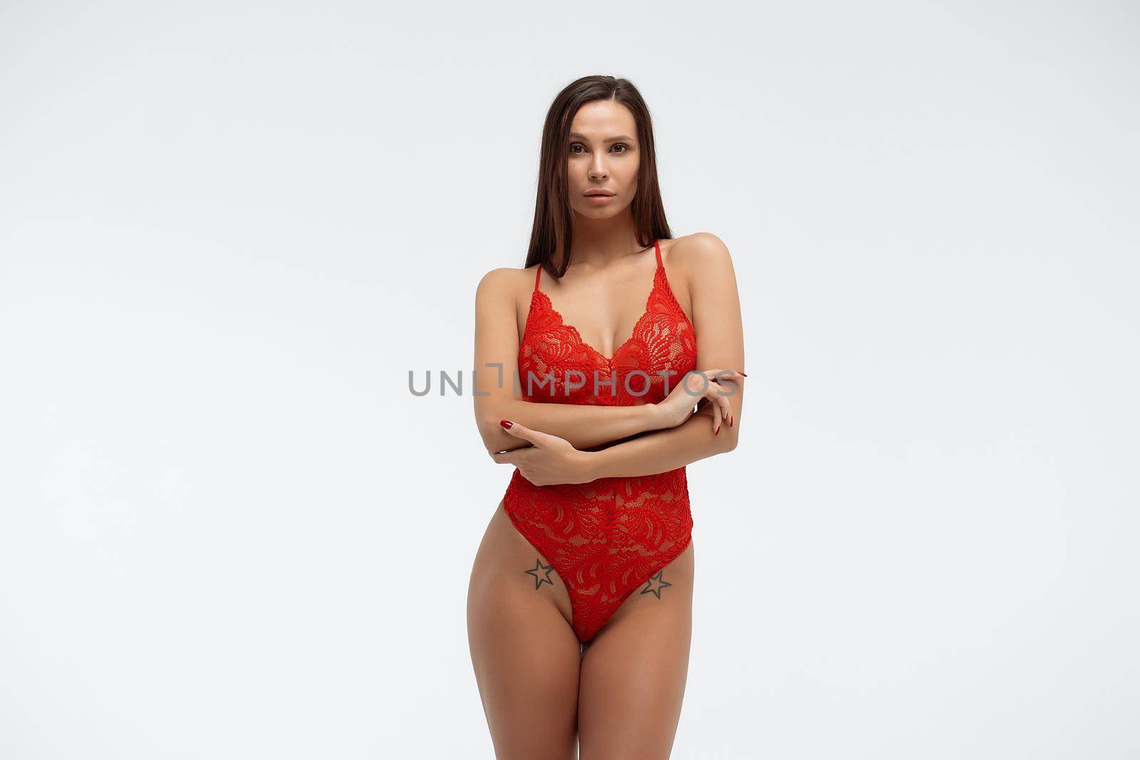 Sexual female with perfect body in red lace bodysuit standing with folded arms on white background and looking at camera